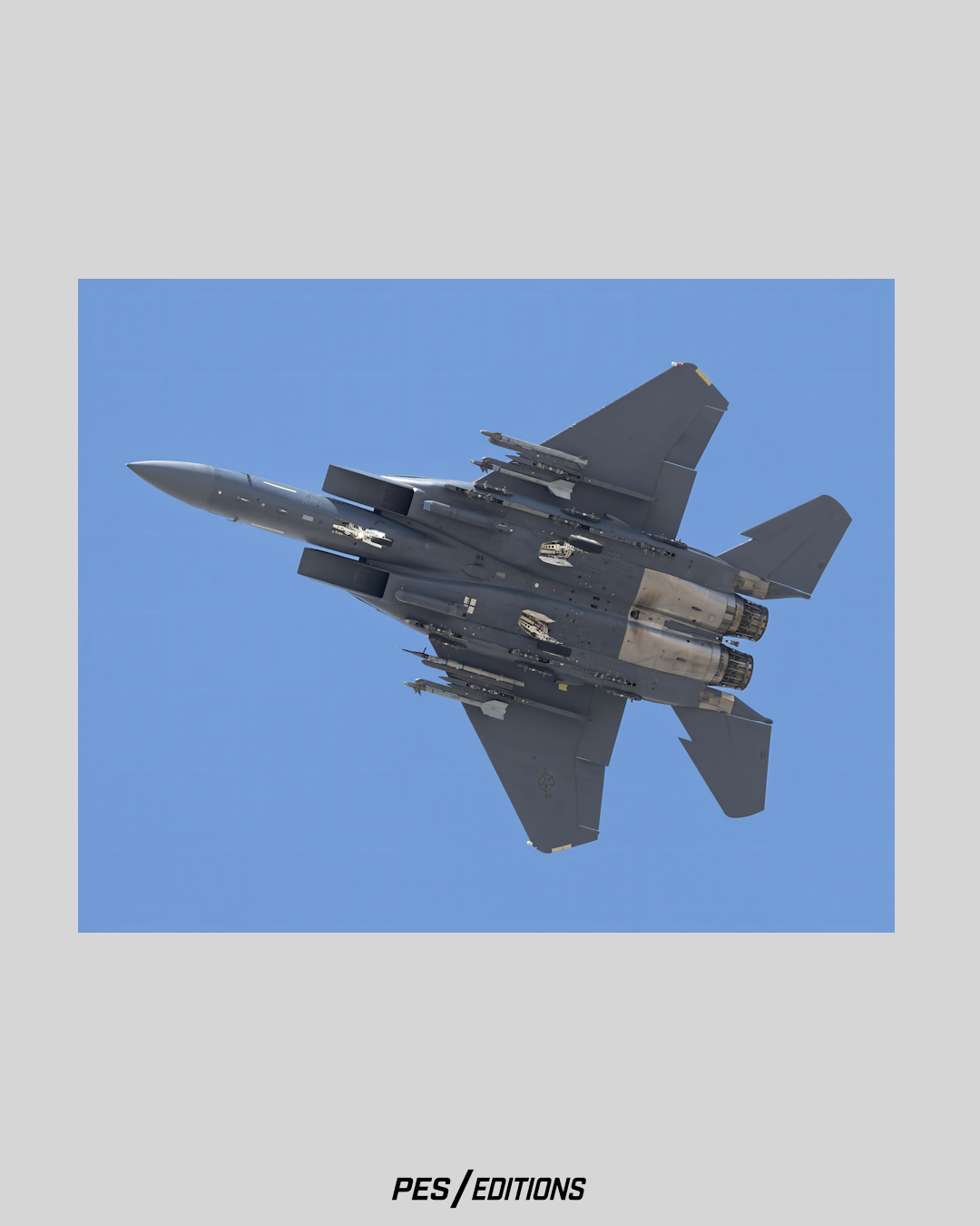 A crisp underside view of a military F-15 Strike Eagle fighter jet banking sharply against a clear blue sky, showing extensive details of its underbelly, wings, dual engines, and weapon rails loaded with missiles.