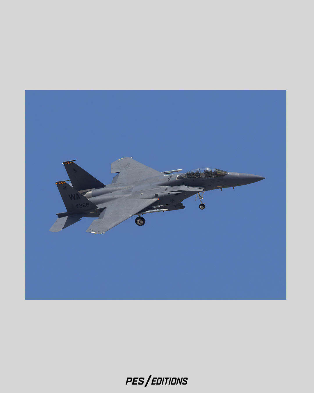 A crisp profile photograph of a two-seat F-15E Strike Eagle fighter jet flying against a clear blue sky, captured from a low angle with its landing gear down as it prepares for approach.