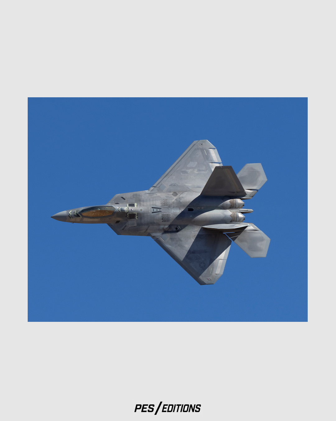 A striking, flat-perspective photograph capturing the underside and full delta-wing silhouette of a U.S. Air Force F-22A Raptor stealth fighter banking sharply against a solid, clear blue sky.